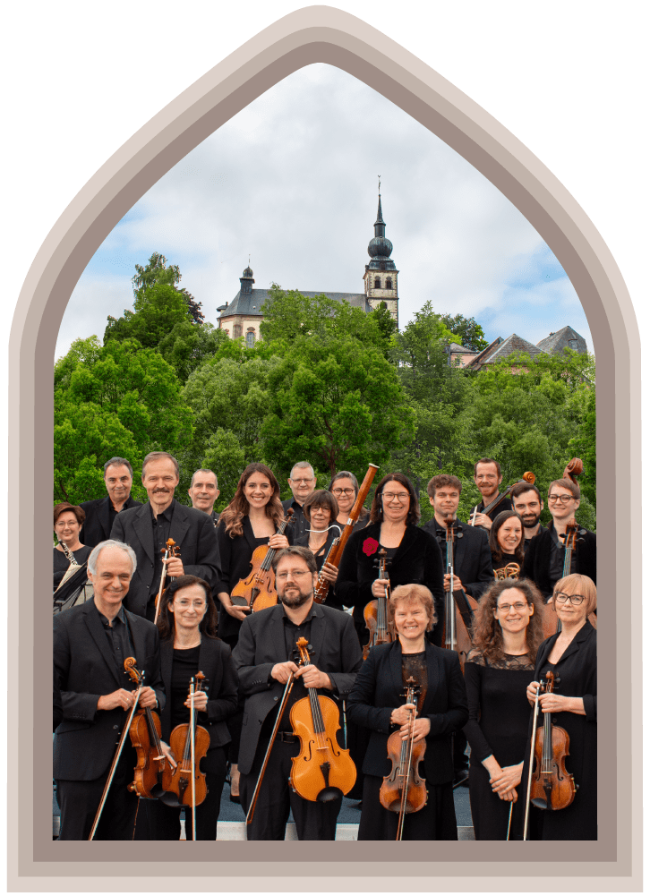 Smiling musicians from the Estro Armonico orchestra stand with their instruments under a cloudy sky with a church in the background.